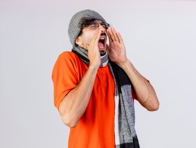 Jeune homme malade portant des lunettes chapeau d'hiver et écharpe en gardant les mains près de la bouche s'apprête à éternuer isolé sur mur blanc