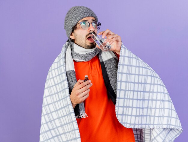 Jeune homme malade de race blanche portant des lunettes chapeau d'hiver et écharpe enveloppé dans un plaid tenant un médicament en verre à la recherche d'un verre d'eau potable isolé sur fond violet