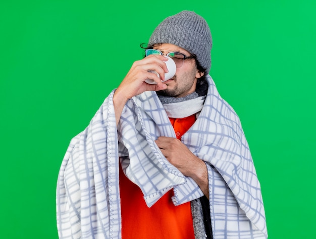 Photo gratuite jeune homme malade de race blanche portant des lunettes chapeau d'hiver et une écharpe enveloppée de plaid saisissant plaid à l'intérieur de la tasse de boire du thé isolé sur le mur vert