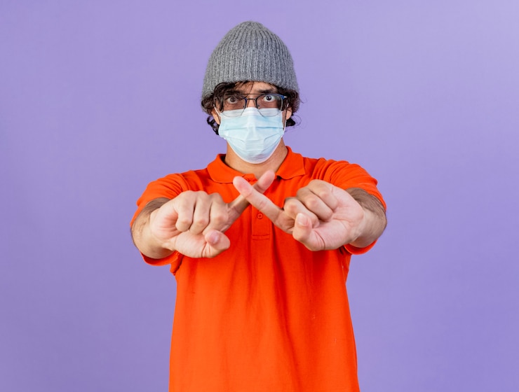Photo gratuit jeune homme malade de race blanche portant des lunettes masque et chapeau d'hiver ne faisant aucun geste isolé sur un mur violet avec espace de copie