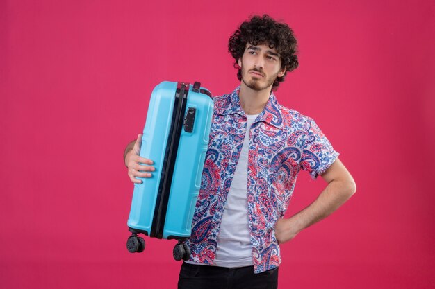 Jeune homme malheureux beau voyageur bouclé tenant la valise avec la main sur la taille sur un mur rose isolé avec espace de copie