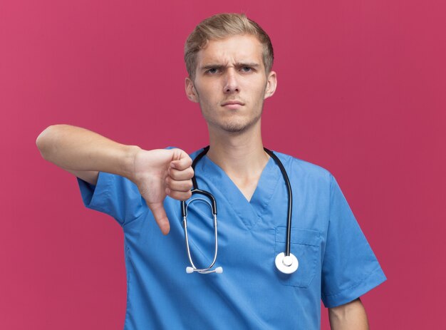 Jeune homme malheureux médecin portant l'uniforme de médecin avec stéthoscope montrant le pouce vers le bas isolé sur un mur rose