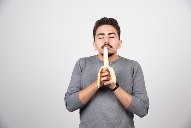 Un jeune homme mange une banane sur un mur blanc.