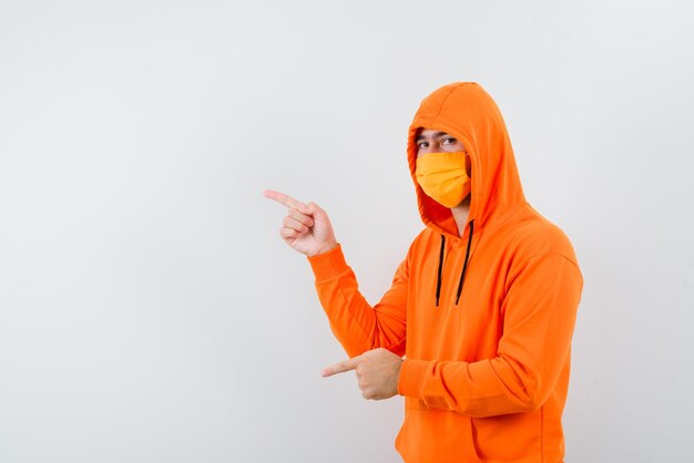 Jeune homme avec un masque montrant la gauche avec ses doigts sur fond blanc