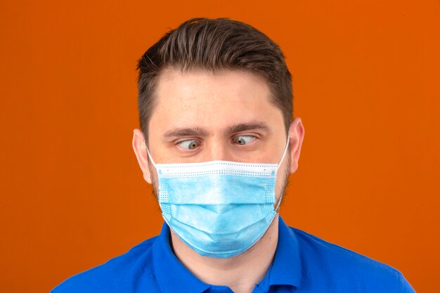 Jeune homme en masque de protection médicale faisant grimace en traversant les yeux sur un mur orange isolé