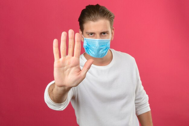 Jeune homme en masque de protection médicale faisant panneau d'arrêt avec la main regardant la caméra avec un visage sérieux debout sur fond rose isolé