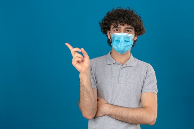 Jeune homme en masque de protection médicale pointant vers le côté regardant la caméra sur fond bleu isolé