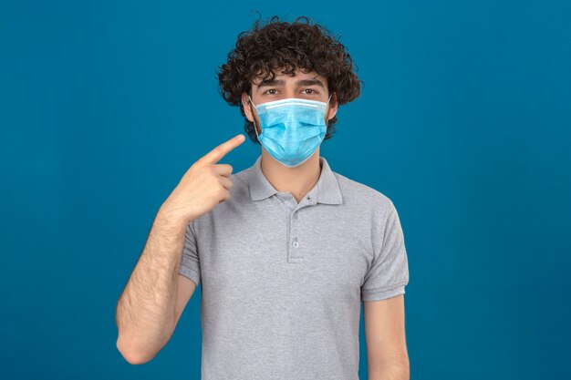 Jeune homme en masque de protection médicale pointant vers son masque regardant la caméra avec sourire sur fond bleu isolé