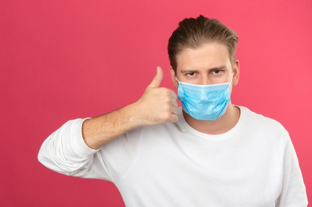 Jeune homme en masque de protection médicale regardant la caméra montrant les pouces vers le haut debout sur fond rose isolé