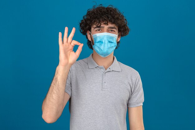 Jeune homme en masque de protection médicale regardant la caméra avec sourire faisant signe ok sur fond bleu isolé