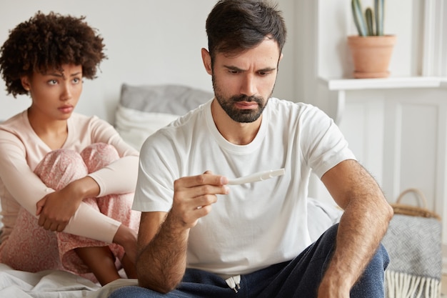 Un jeune homme mécontent et mal rasé tient un test de grossesse, se sent stressé par les résultats positifs, attend un bébé indésirable. Une femme mécontente est assise derrière.