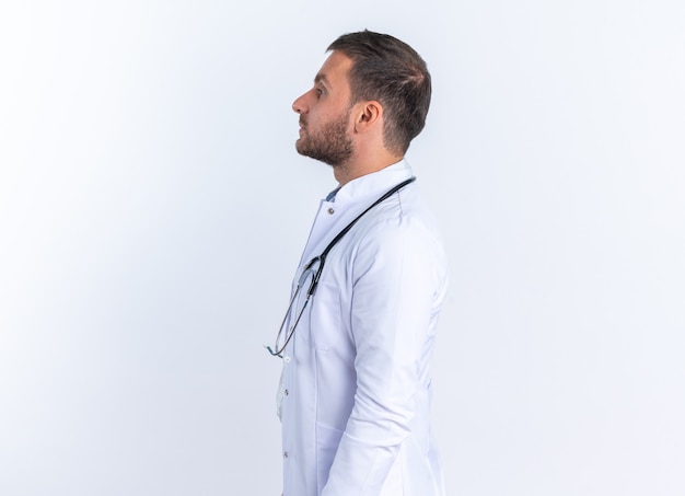 Jeune homme médecin en blouse blanche et avec stéthoscope autour du cou, l'air confiant, debout sur le mur blanc