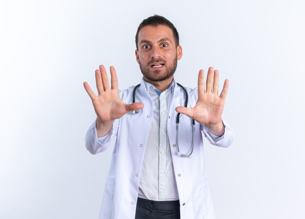 Jeune homme médecin en blouse blanche et avec stéthoscope autour du cou inquiet avec les mains comme disant de ne pas s'approcher debout sur un mur blanc