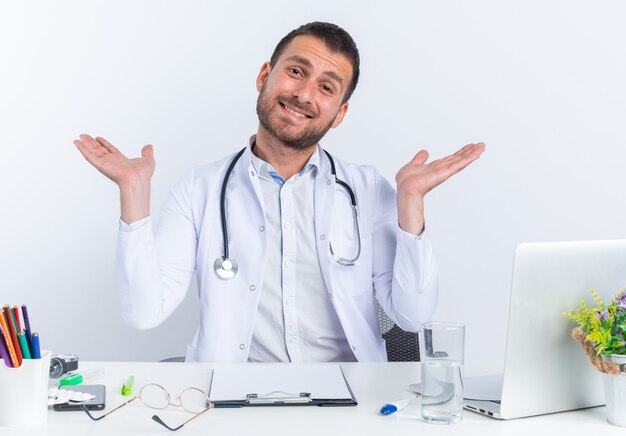 Jeune homme médecin en blouse blanche et avec stéthoscope regardant à l'avant un sourire heureux et joyeux écartant largement les bras sur les côtés assis à la table avec un ordinateur portable sur un mur blanc