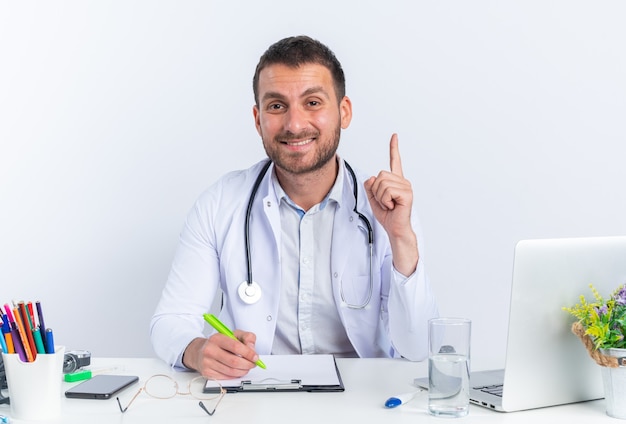 Jeune homme médecin en blouse blanche et avec stéthoscope souriant écrit confiant montrant l'index ayant une bonne idée assis à la table avec un ordinateur portable sur un mur blanc