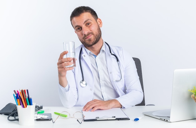Jeune homme médecin en blouse blanche et avec stéthoscope tenant un verre d'eau heureux et positif souriant confiant assis à la table avec un ordinateur portable sur un mur blanc