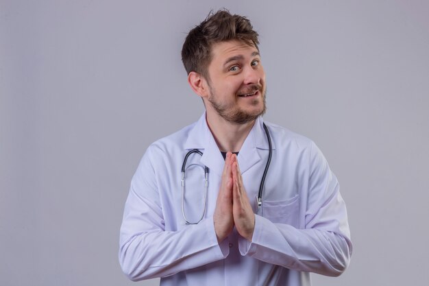 Jeune homme médecin heureux et positif portant blouse blanche et stéthoscope main dans la main en geste de namaste