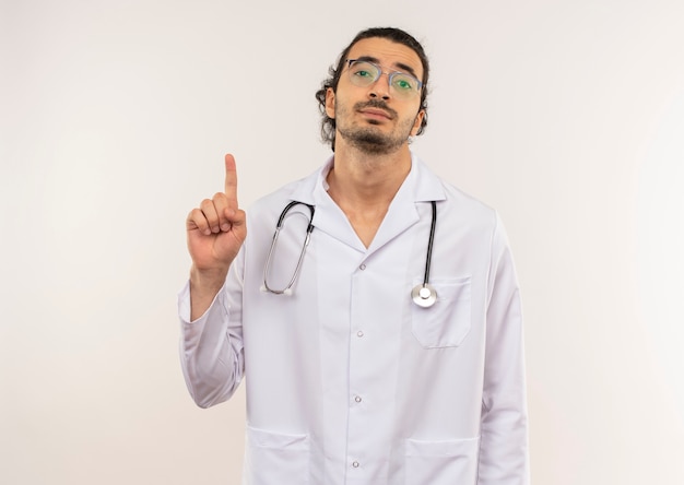 Jeune homme médecin avec des lunettes optiques portant une robe blanche avec stéthoscope pointe vers le haut