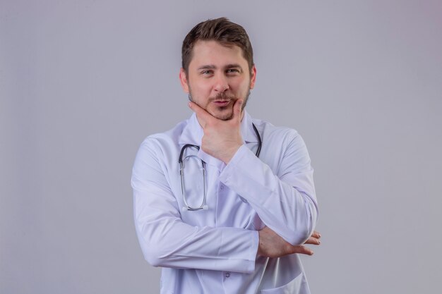 Jeune homme médecin portant blouse blanche et stéthoscope garde la main sous le menton, pense à quelque chose d'agréable