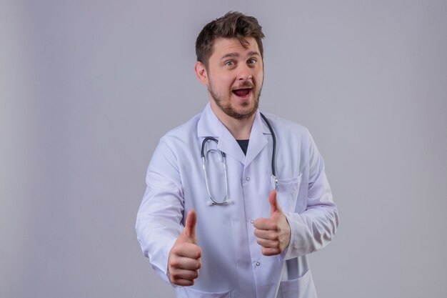 Jeune homme médecin portant blouse blanche et stéthoscope montrant le geste du pouce en l'air avec un visage heureux 1