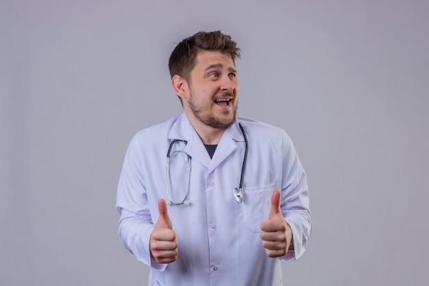 Jeune homme médecin portant blouse blanche et stéthoscope montrant le geste du pouce en l'air avec un visage heureux 3