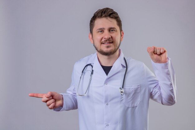 Jeune homme médecin portant blouse blanche et stéthoscope pointant le doigt sur le côté et levant le poing avec sourire sur le visage