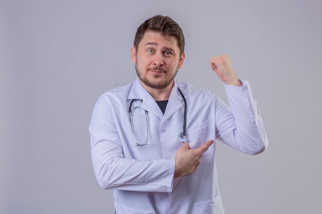 Jeune homme médecin portant blouse blanche et stéthoscope pointant sur ses biceps, expression de force, concept gagnant