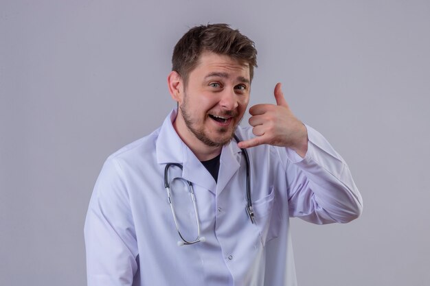 Jeune homme médecin portant blouse blanche et stéthoscope souriant faisant parler au téléphone geste appelez-moi signe