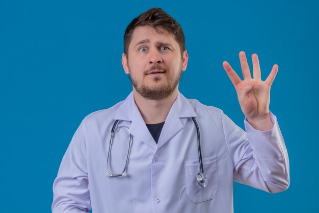 Jeune homme médecin portant blouse blanche et stéthoscope avec sourire montrant le numéro trois avec les doigts sur fond bleu isolé