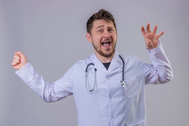 Jeune homme médecin portant blouse blanche et stéthoscope surpris avec un visage heureux 1