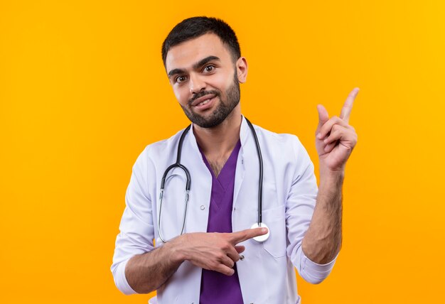 Jeune homme médecin portant une robe médicale stéthoscope indique une direction différente sur un mur jaune isolé