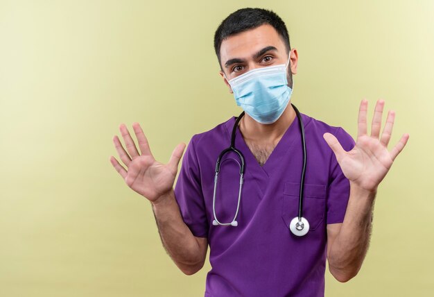 Jeune homme médecin portant des vêtements de chirurgien violet et masque médical stéthoscope levant les mains sur un mur vert isolé