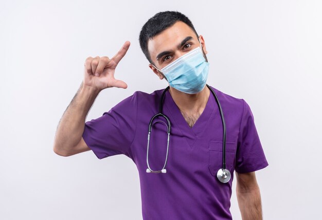 Jeune homme médecin portant des vêtements de chirurgien violet et masque médical stéthoscope montrant la taille sur un mur blanc isolé