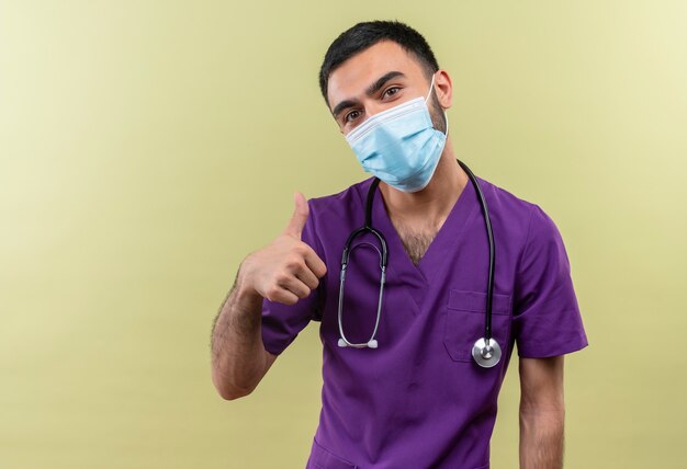 Jeune homme médecin portant des vêtements de chirurgien violet et un masque médical stéthoscope son pouce vers le haut sur un mur vert isolé