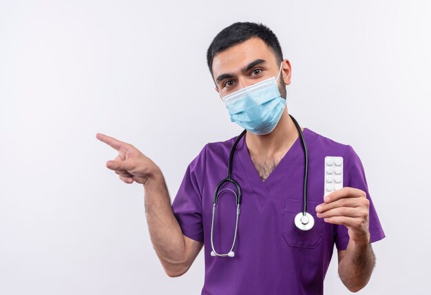 Jeune homme médecin portant des vêtements de chirurgien violet et un masque médical stéthoscope tenant des pilules et des points à côté sur un mur blanc isolé