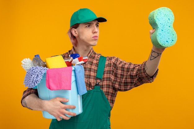 Jeune homme de ménage en combinaison chemise à carreaux et casquette tenant une éponge et un seau avec des outils de nettoyage regardant de côté avec un visage sérieux prêt pour le nettoyage debout sur orange