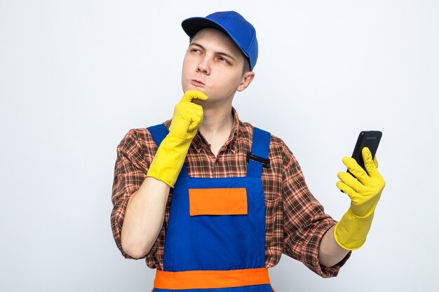 Jeune homme de ménage portant un uniforme et une casquette avec des gants tenant un téléphone isolé sur un mur blanc