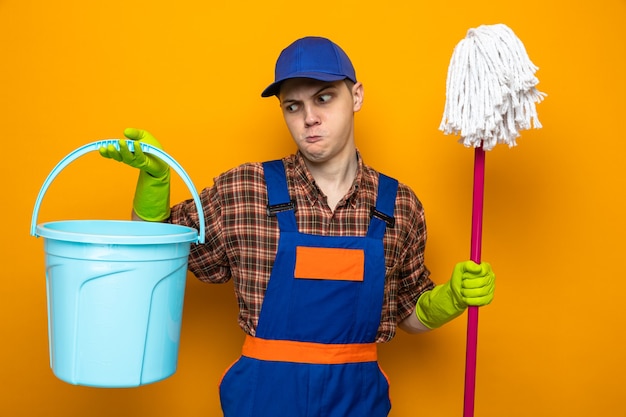 Jeune homme de ménage portant un uniforme et une casquette avec des gants tenant une vadrouille et un seau isolés sur un mur orange