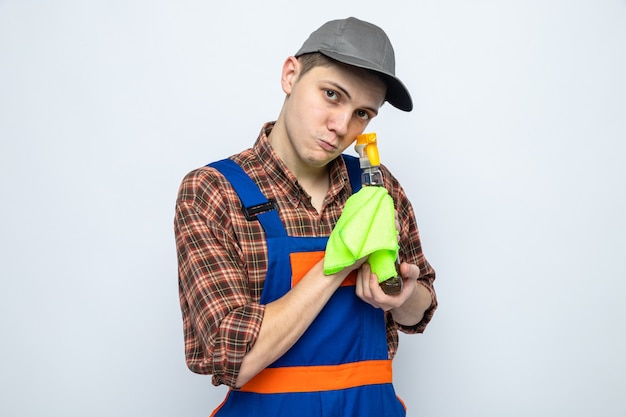 Jeune homme de ménage portant un uniforme et une casquette tenant un chiffon avec un agent de nettoyage isolé sur un mur blanc