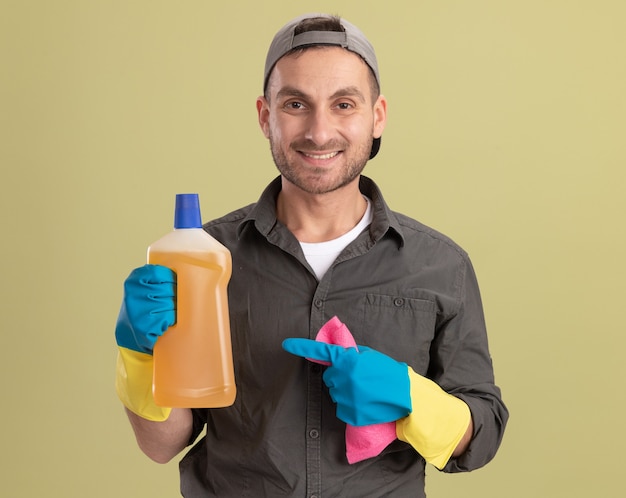 Jeune homme de ménage portant des vêtements décontractés et capuchon dans des gants en caoutchouc tenant une bouteille avec des produits de nettoyage et un chiffon à sourire heureux et positif debout sur le mur vert