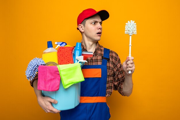 Jeune homme de ménage en uniforme et casquette tenant un seau d'outils de nettoyage et regardant une brosse dans sa main isolée sur un mur orange