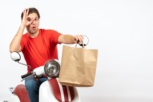 Jeune Homme De Messagerie Confiant En Uniforme Rouge Assis Sur Un Scooter Donnant Un Sac En Papier En Regardant Quelque Chose Sur Le Mur Blanc