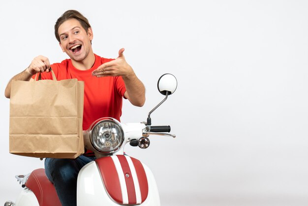 Jeune homme de messagerie souriant en uniforme rouge assis sur un sac de papier de pointage scooter sur mur blanc