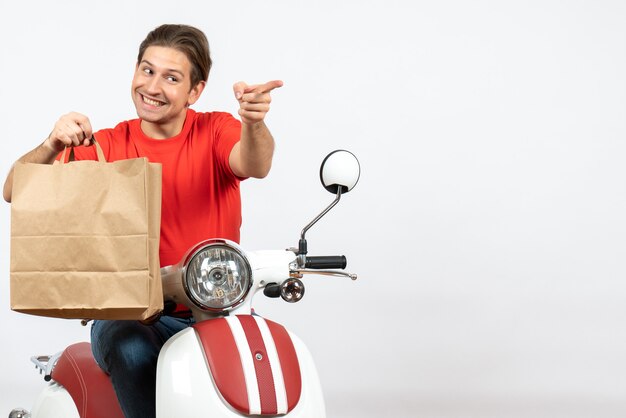 Jeune homme de messagerie souriant en uniforme rouge assis sur un scooter tenant un sac en papier pointant vers l'avant sur un mur blanc