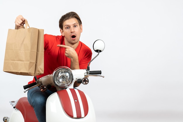 Jeune homme de messagerie surpris en uniforme rouge assis sur un sac en papier pointant scooter sur mur jaune