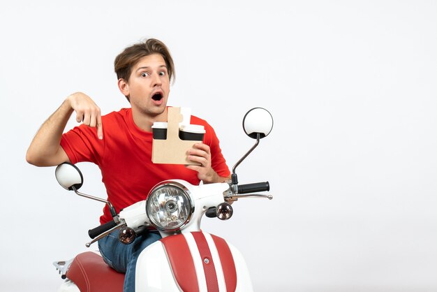 Jeune homme de messagerie surpris en uniforme rouge assis sur un scooter tenant une tasse de papier pointant vers le bas sur le mur jaune