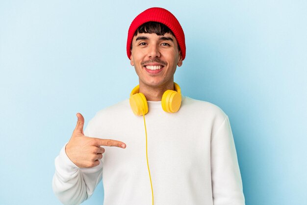 Jeune homme métis écoutant de la musique isolé sur fond bleu personne pointant à la main vers un espace de copie de chemise, fier et confiant