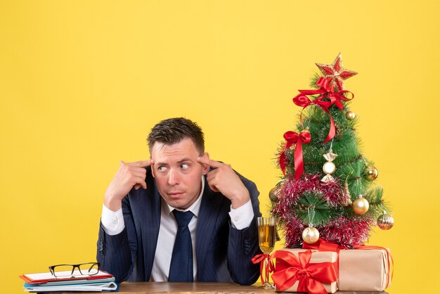 Jeune homme mettant les doigts à son temple assis à la table près de l'arbre de Noël et des cadeaux sur jaune