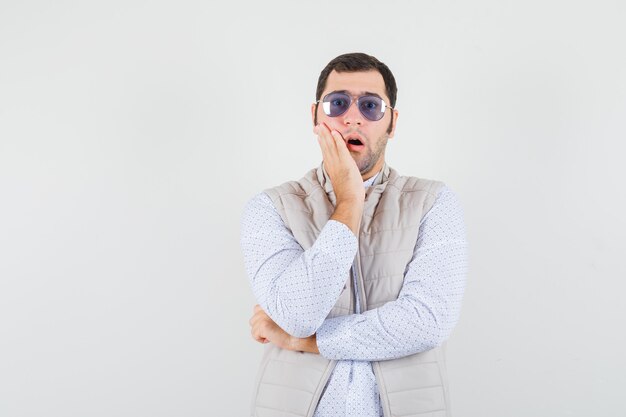 Jeune homme mettant des lunettes tout en mettant la main près de la bouche en veste beige et casquette et à la vue de face surprise.