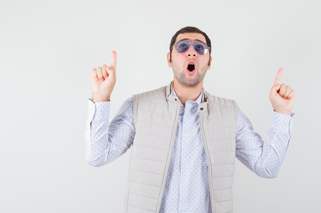 Jeune homme mettant des lunettes tout en pointant vers le haut avec l'index en veste beige et casquette et à la surprise, vue de face.